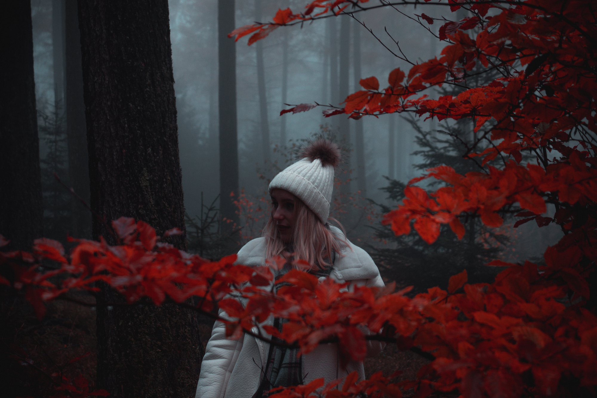 a woman in a white hat and white coat standing in a forest with red leaves