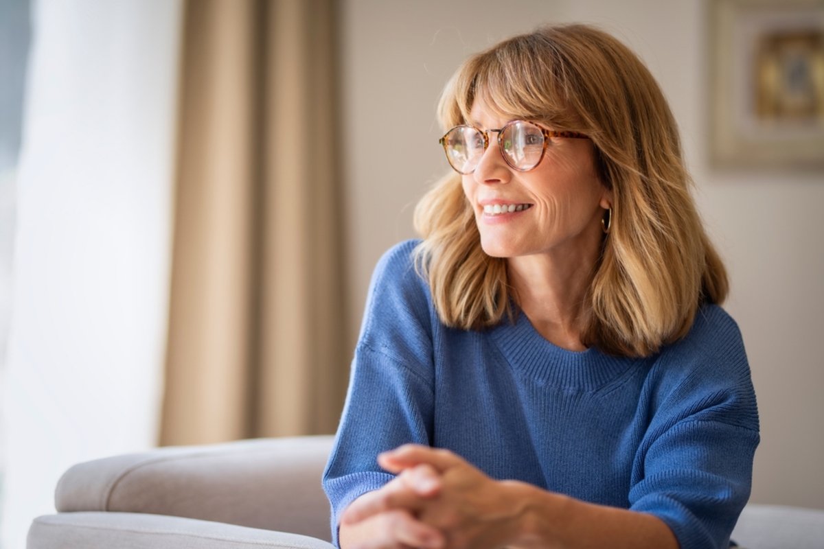 Lächelnde Frau mit Brille und blauem Pullover sitzt am Fenster, Lichtreflexe auf den Brillengläsern | Luminar Blog