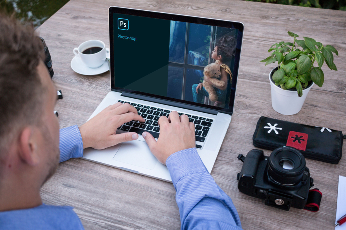 Photoshop-Ladebildschirm auf Laptop neben Kamera, Kaffeetasse und Topfpflanze am Holztisch | Luminar Blog